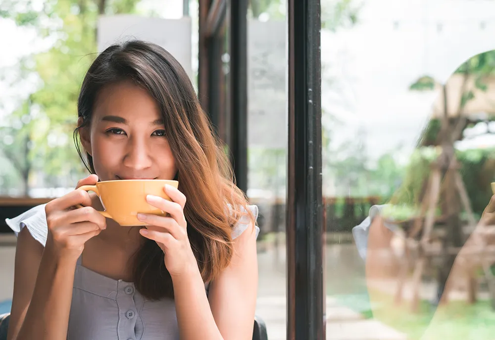 cheerful-asian-young-woman-drinking-warm-coffee-tea-enjoying-it-while-sitting-cafe